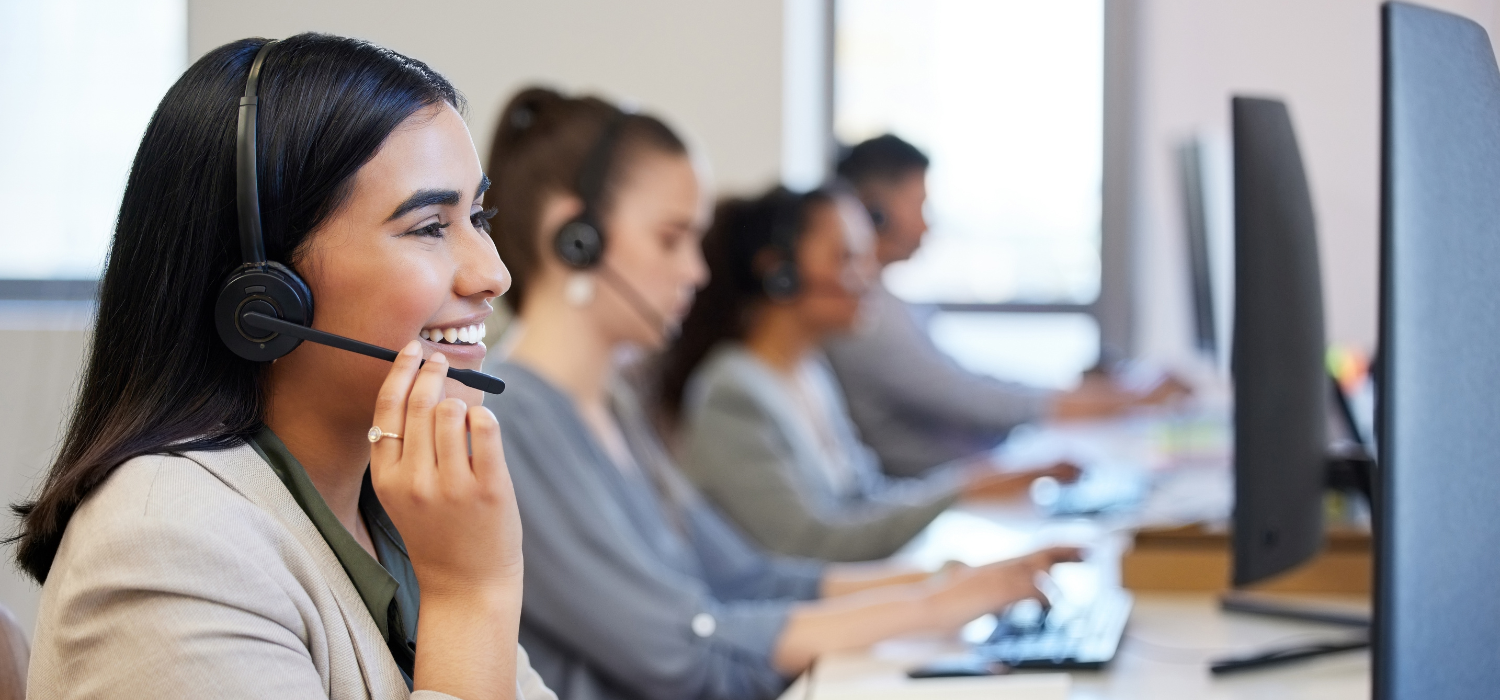 A smiling customer service representative wearing a headset, working at a computer in a call centre alongside colleagues.