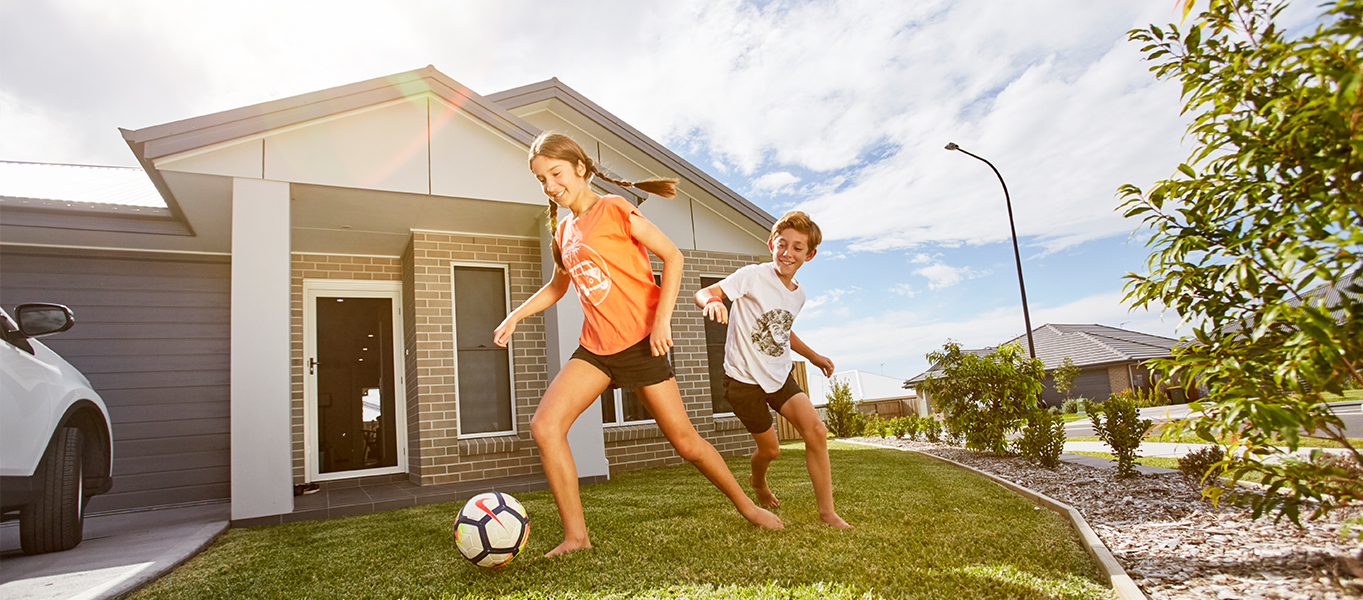 Two children playing soccer on the front lawn of a modern brick home in a residential neighbourhood.