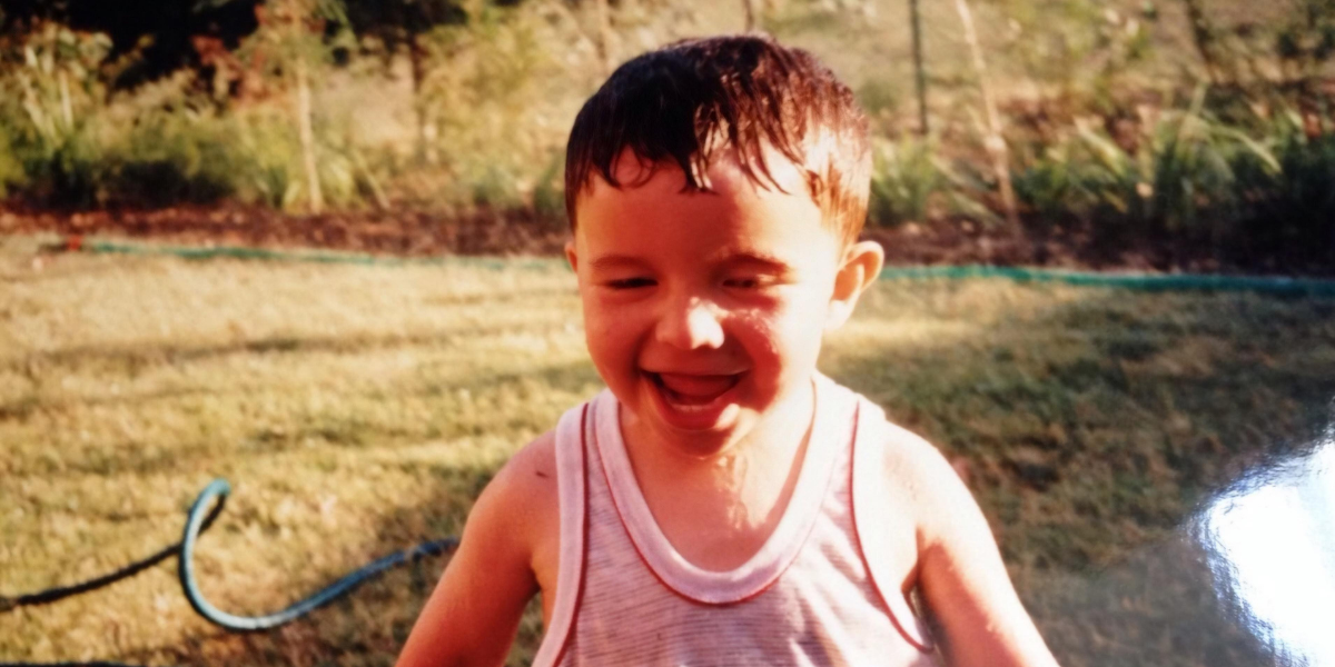 Simon's son as a toddler smiling in the backyard. There is lots of grass and very small bushes along the fence in the background.