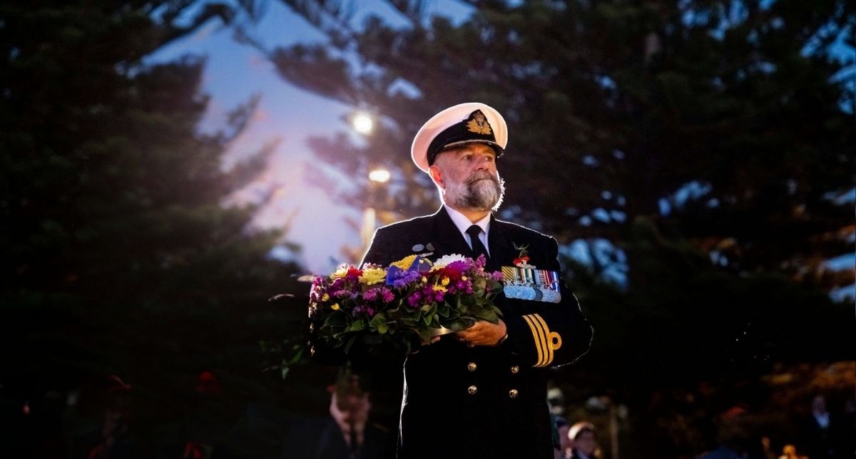 Commander Simon Kelly holding a wreath of flowers at a dawn service in 2025. There are tall pine trees in the background and the sun is slowly rising behind him.