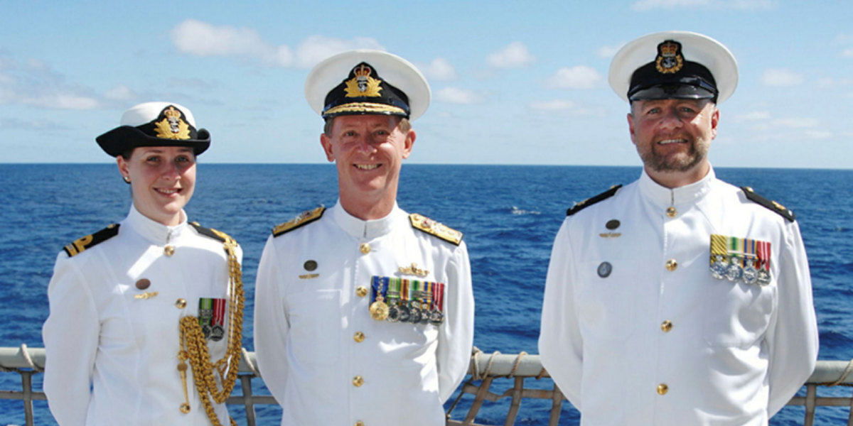 Simon (far right) as a Warrant Officer accompanying the Commander of the Australian Fleet and Flag Lieutenant. They are on the open deck of a large Navy boat in the sun. There is blue sky and water behind them. 