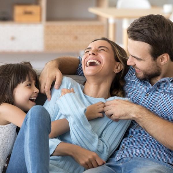 A happy family of three laughing together on a sofa in a bright living room.