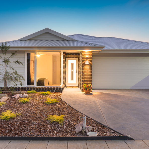 A modern single-storey brick home with a double garage, illuminated entrance, and landscaped front garden at dusk.
