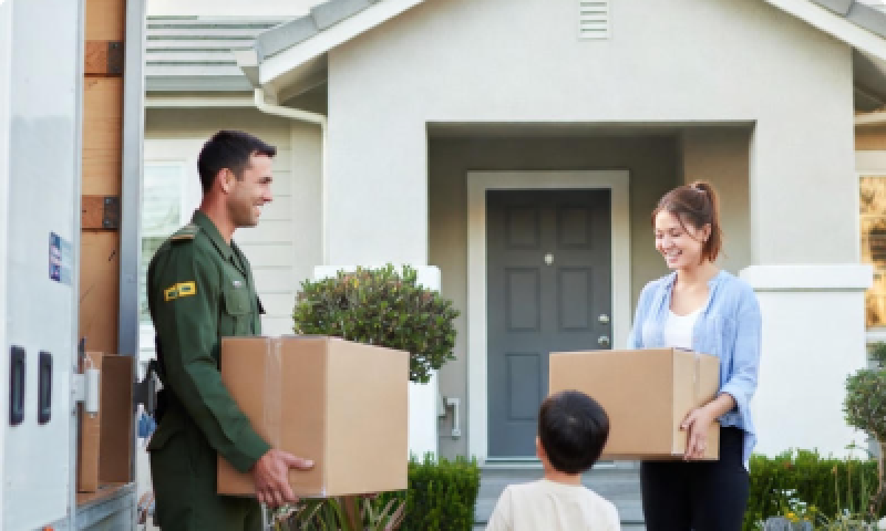 Defence member and partner carry moving boxes towards a house with their child in the foreground.