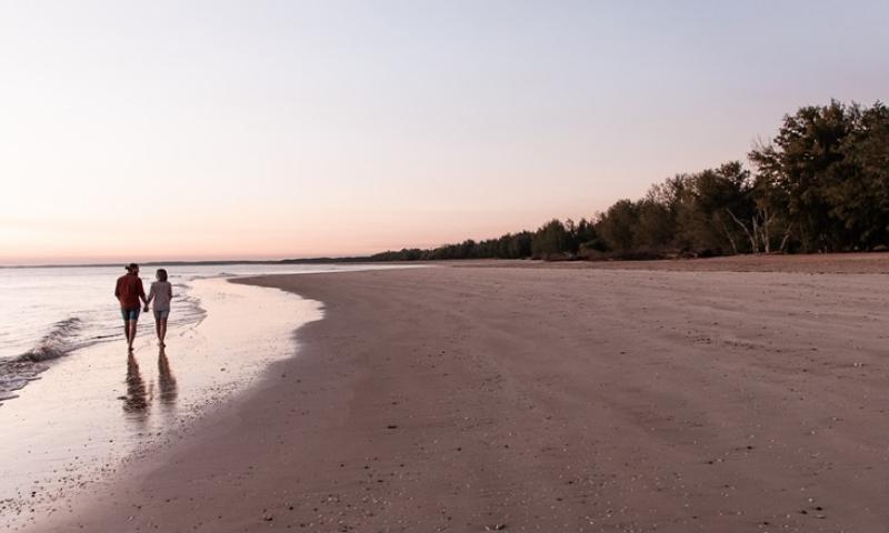 A couple walking hand-in-hand along a quiet beach at sunset, with calm water and a tree-lined shore.