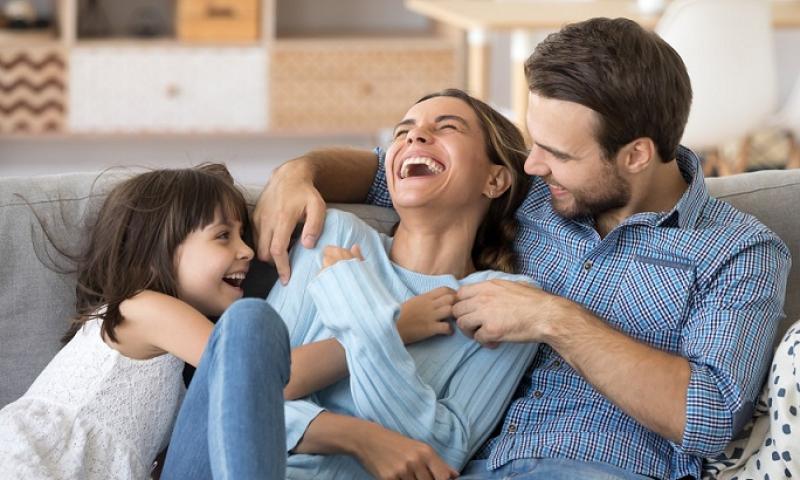 A happy family of three laughing together on a sofa in a bright living room.