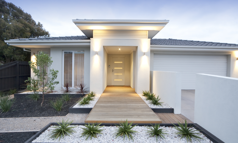 A modern white single-storey home with a timber deck walkway and contemporary landscaping at dusk.